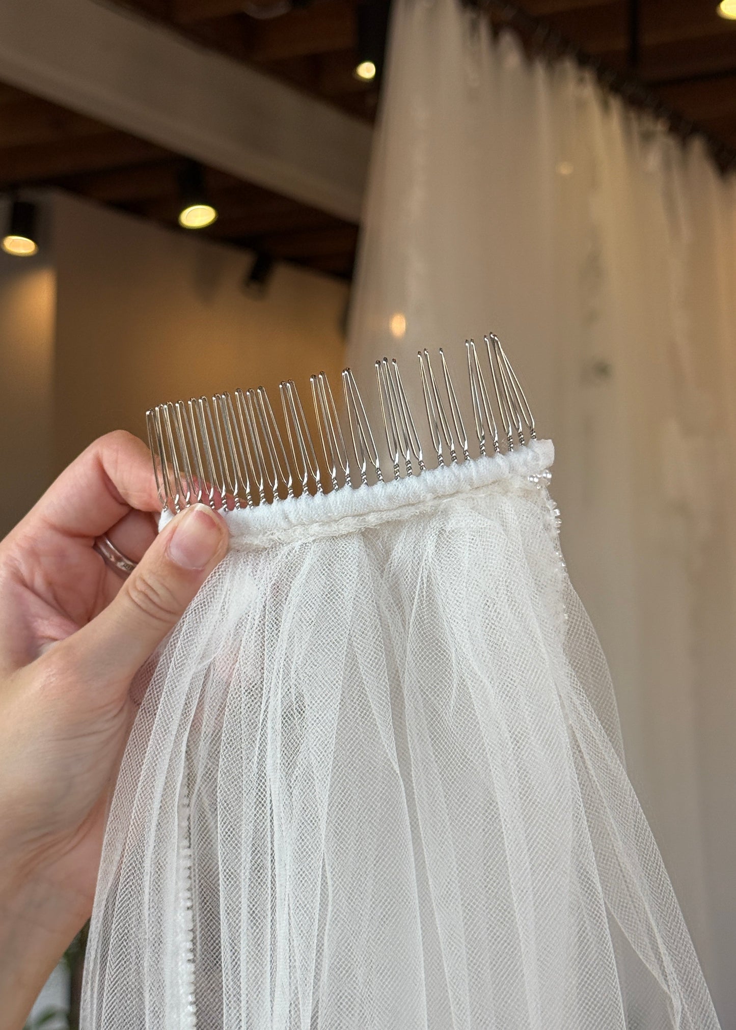 Hand holding a white wedding veil with a clear comb, blurred indoor background