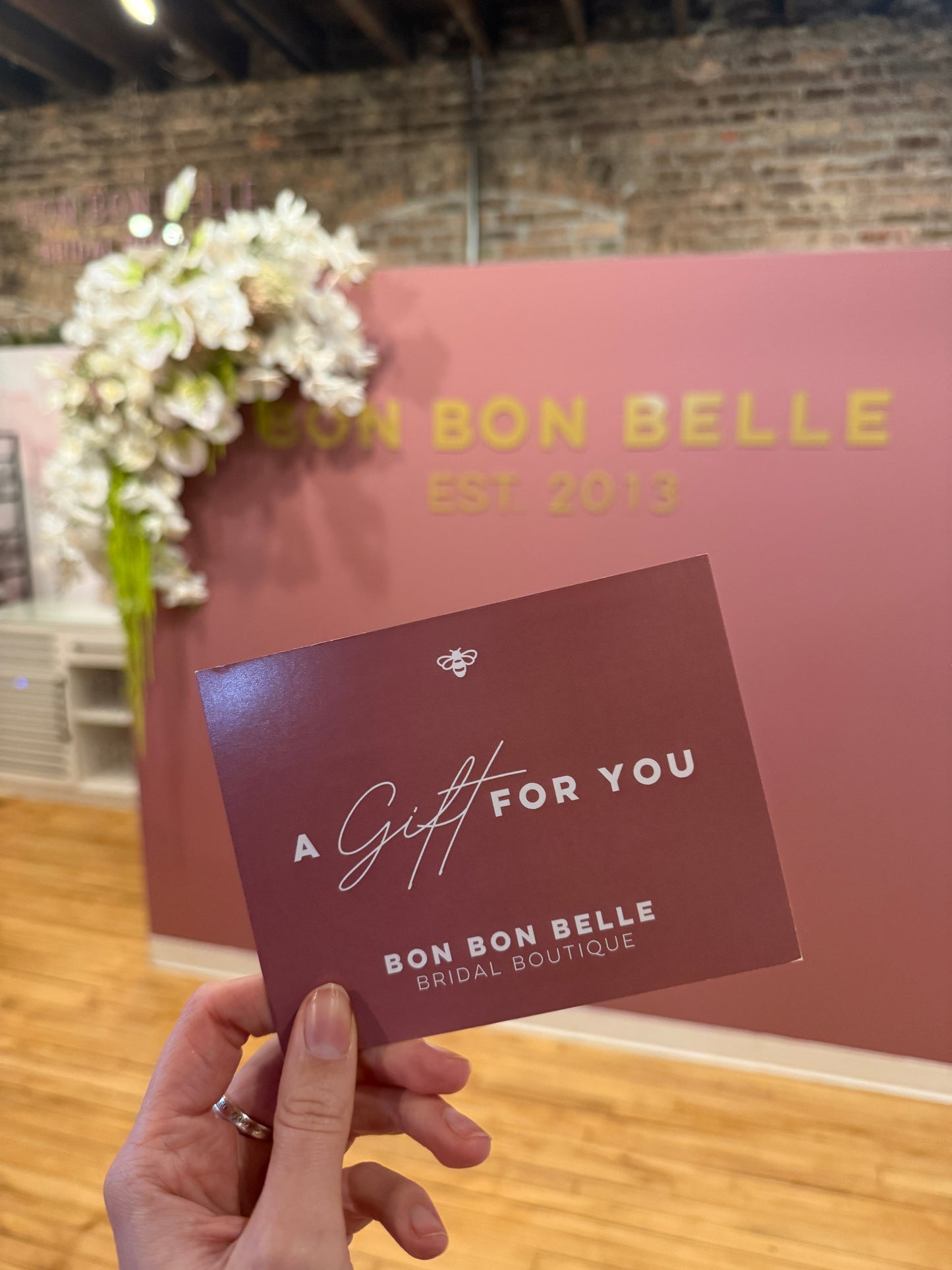 Hand holding a 'Bon Bon Belle' gift card in front of a pink wall with floral decorations.