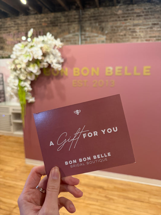 Hand holding a 'Bon Bon Belle' gift card in front of a pink wall with floral decorations.