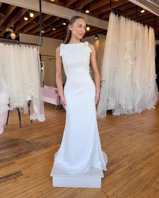 Woman in a white wedding dress standing on a wooden floor with lace curtains in the background.