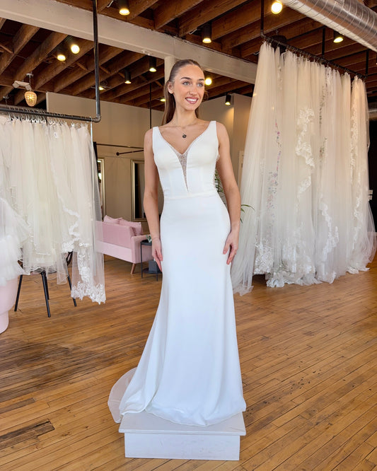 Model wearing a white wedding dress in a bridal shop with lace curtains in the background.